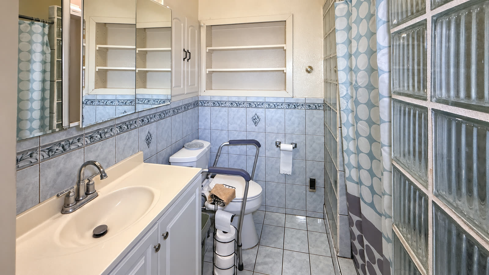 Downstairs bath with a vanity and mirrored cabinet, a shelved wall niche, blue-gray tile wainscot, a glass-block shower wall and a tile floor.