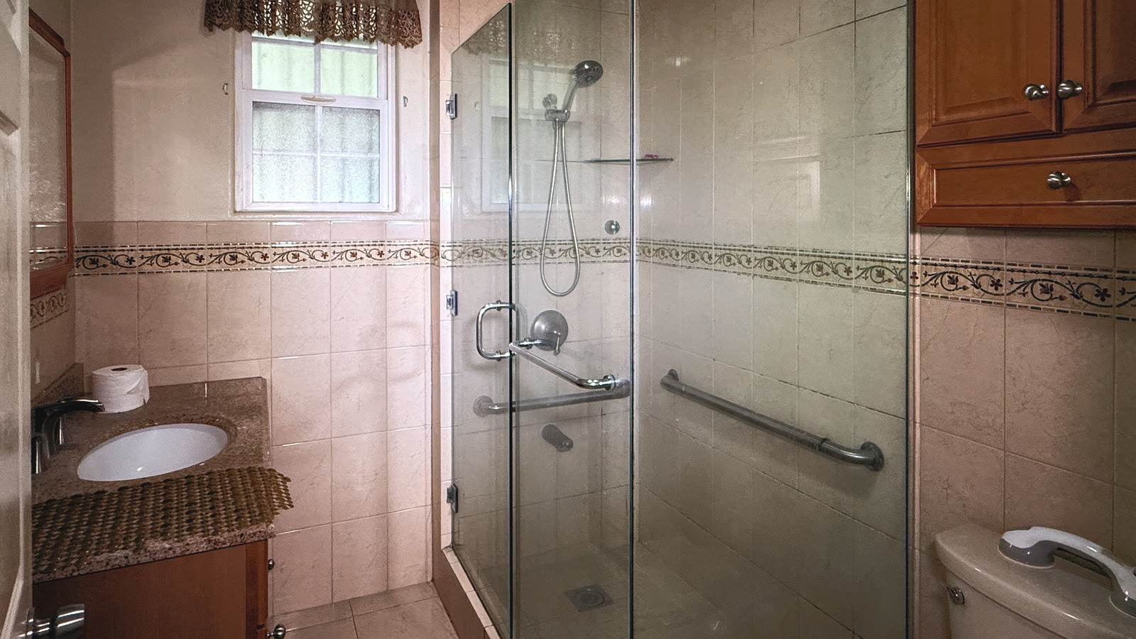 Upstairs bath with a granite vanity top over a dark cabinet, cream wall tile with a floral border, a frameless glass shower with a handheld head and a grab bar.