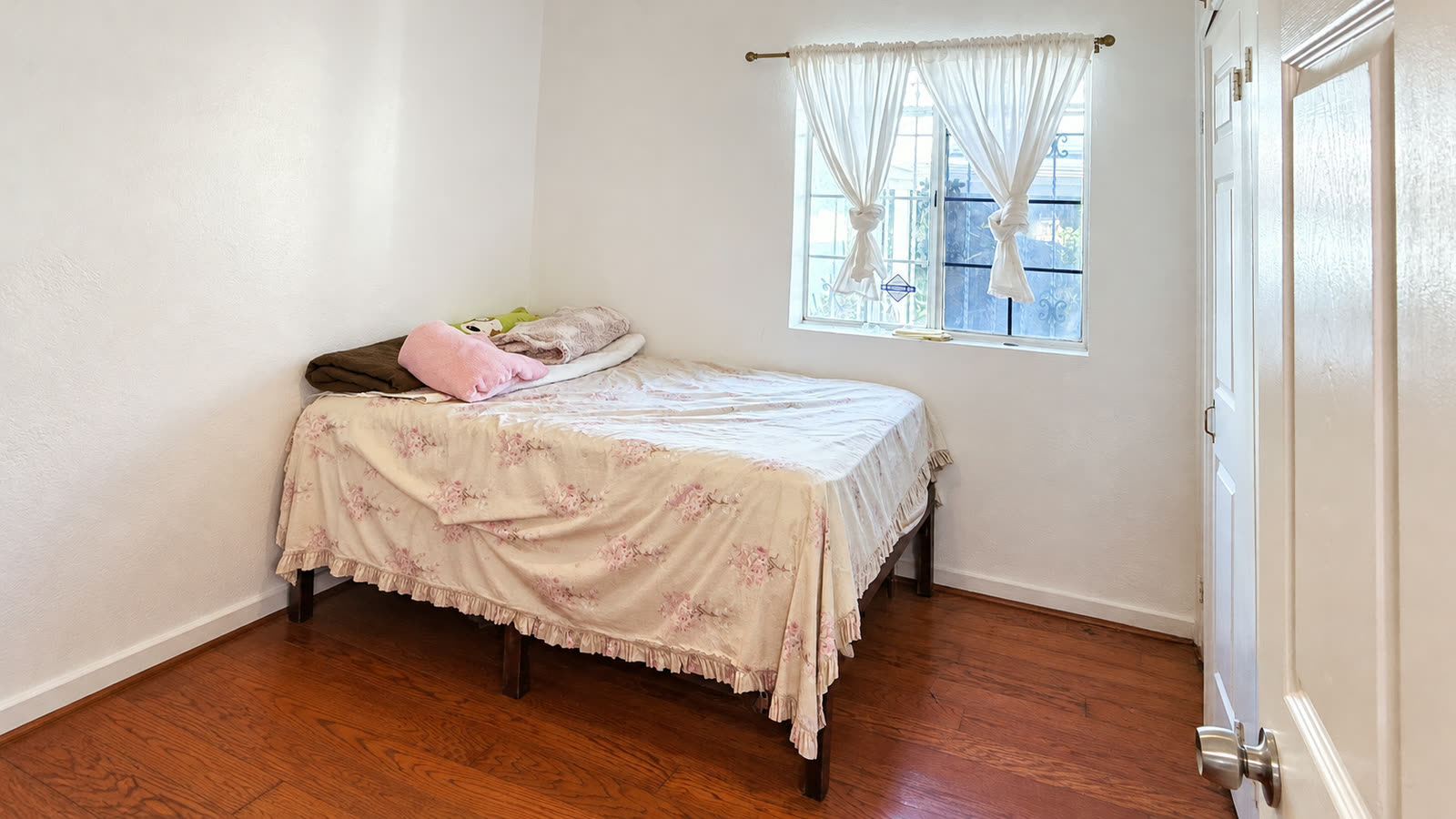 Downstairs bedroom with white walls, a window with sheer curtains, a white six-panel door and dark wood laminate flooring.