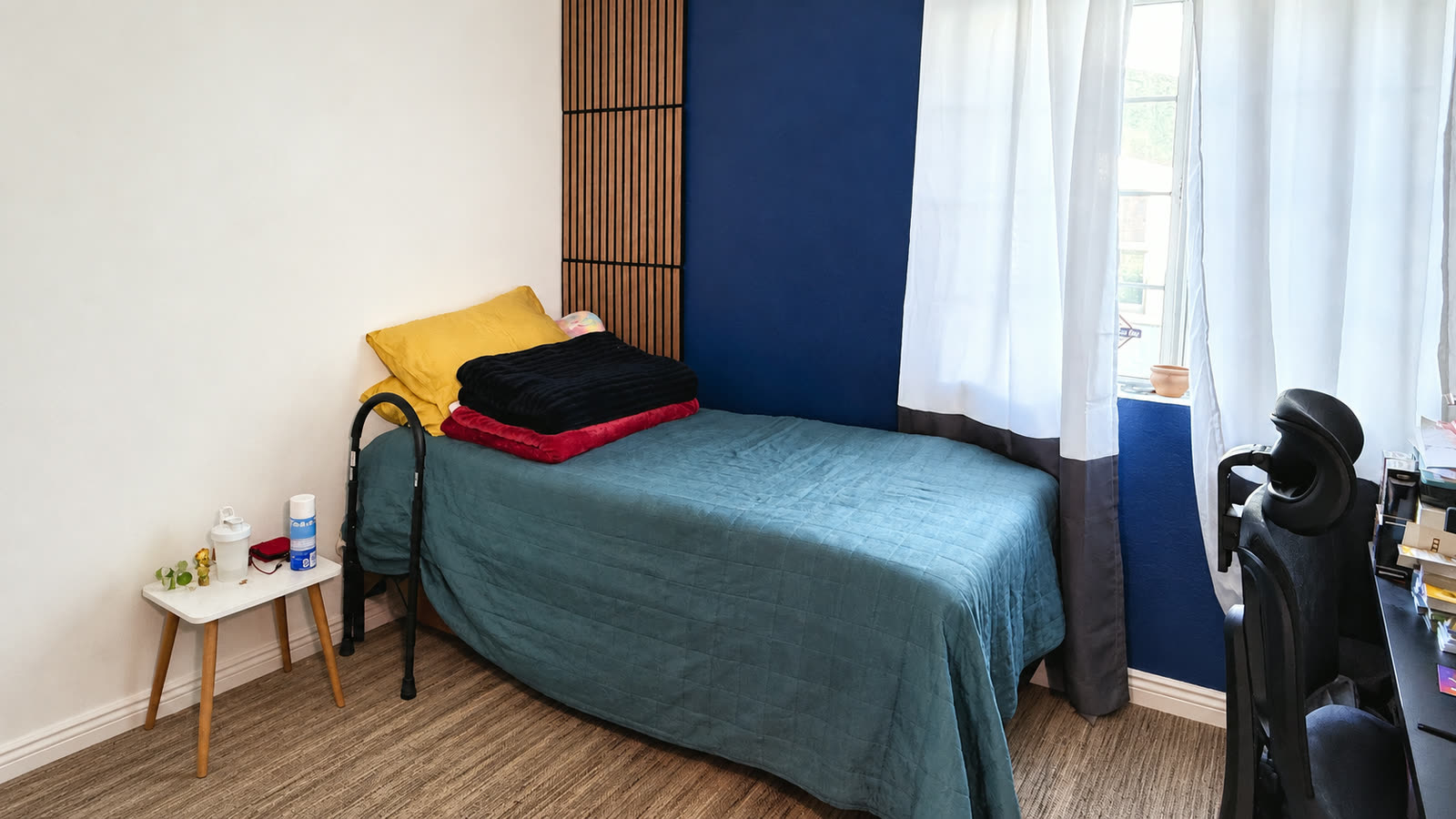 Downstairs bedroom with a vertical wood-slat accent panel and a navy wall behind the bed, and a window with white curtains.