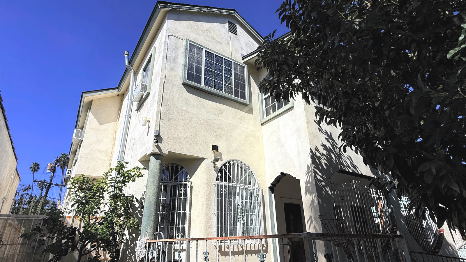 Looking up at the two-story stucco elevation: first-floor arched windows with white iron grilles beside a green-gray column, an arched recess with a dark door, and a large multi-pane upper window.