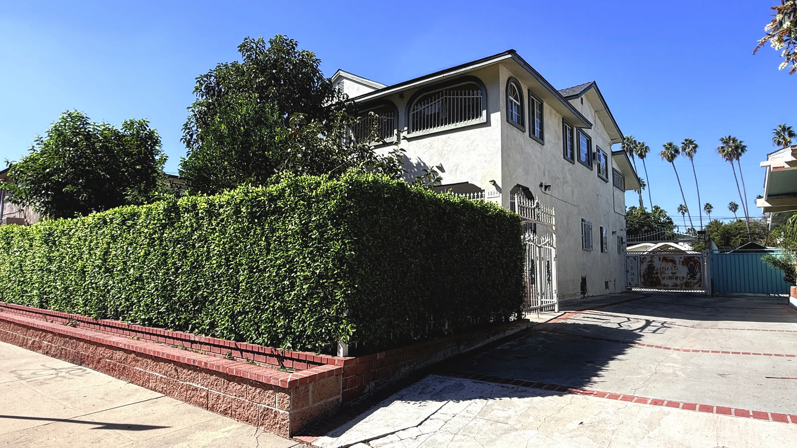 1819 S Norton Ave from the sidewalk: a two-story stucco house with arched upper windows behind white wrought-iron grilles, rising behind a tall clipped hedge on a brick-capped planter wall, with a white iron entry gate at the end of the hedge.