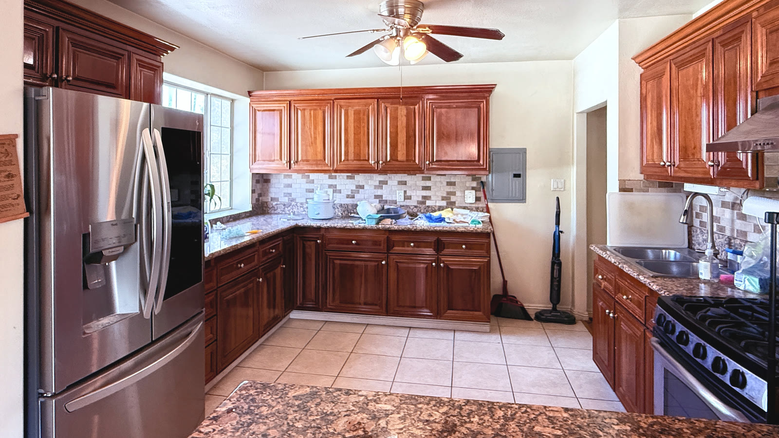 Kitchen from the peninsula: raised-panel cabinets, an L-shaped granite counter under a window, a brick-pattern backsplash and a ceiling fan.