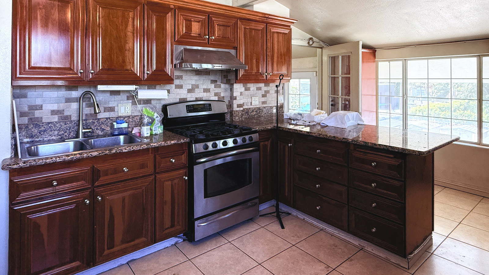 The kitchen: reddish-brown raised-panel cabinets, a tan, gray and cream brick-pattern tile backsplash, a stainless-look gas range under a hood, a double sink and granite counters wrapping into an L-shaped peninsula.