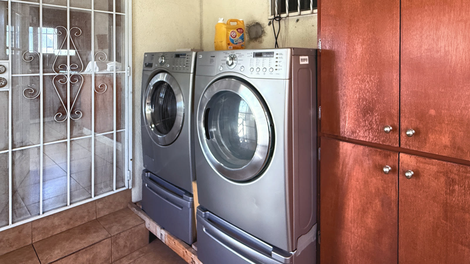 Upstairs laundry area with front-loading machines on pedestals, a barred window above and a tall cabinet at right.