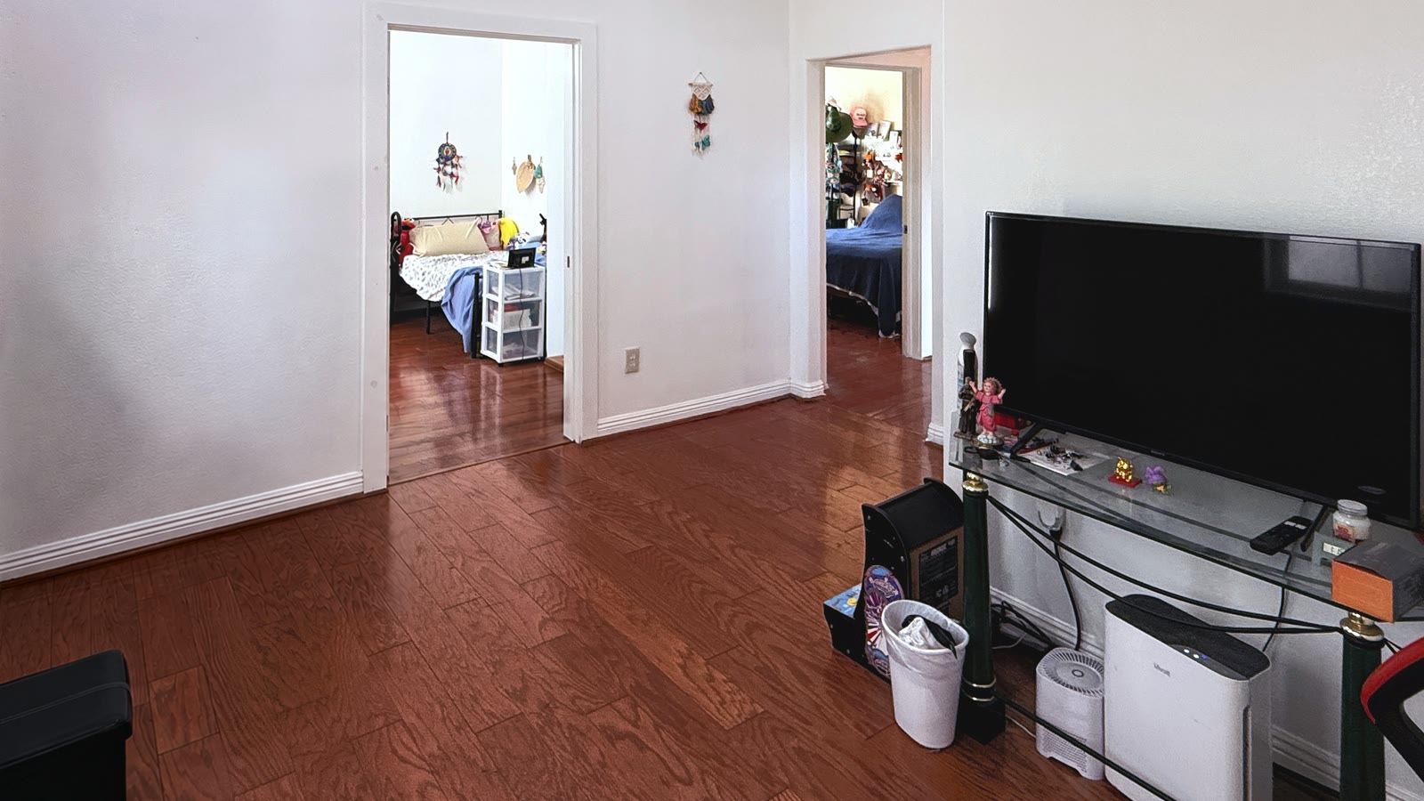 Downstairs living room with wood laminate flooring and white walls, two open doorways on the far wall leading to bedrooms, and a TV on a glass-top stand.