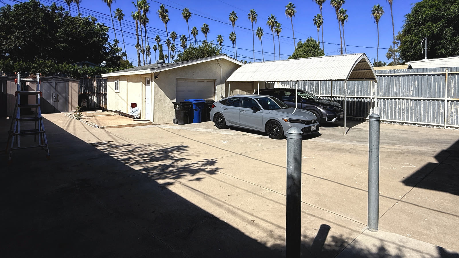 The rear court: a concrete parking area in front of the detached stucco garage with a wide white overhead door, and an arched carport canopy on thin poles beside it.