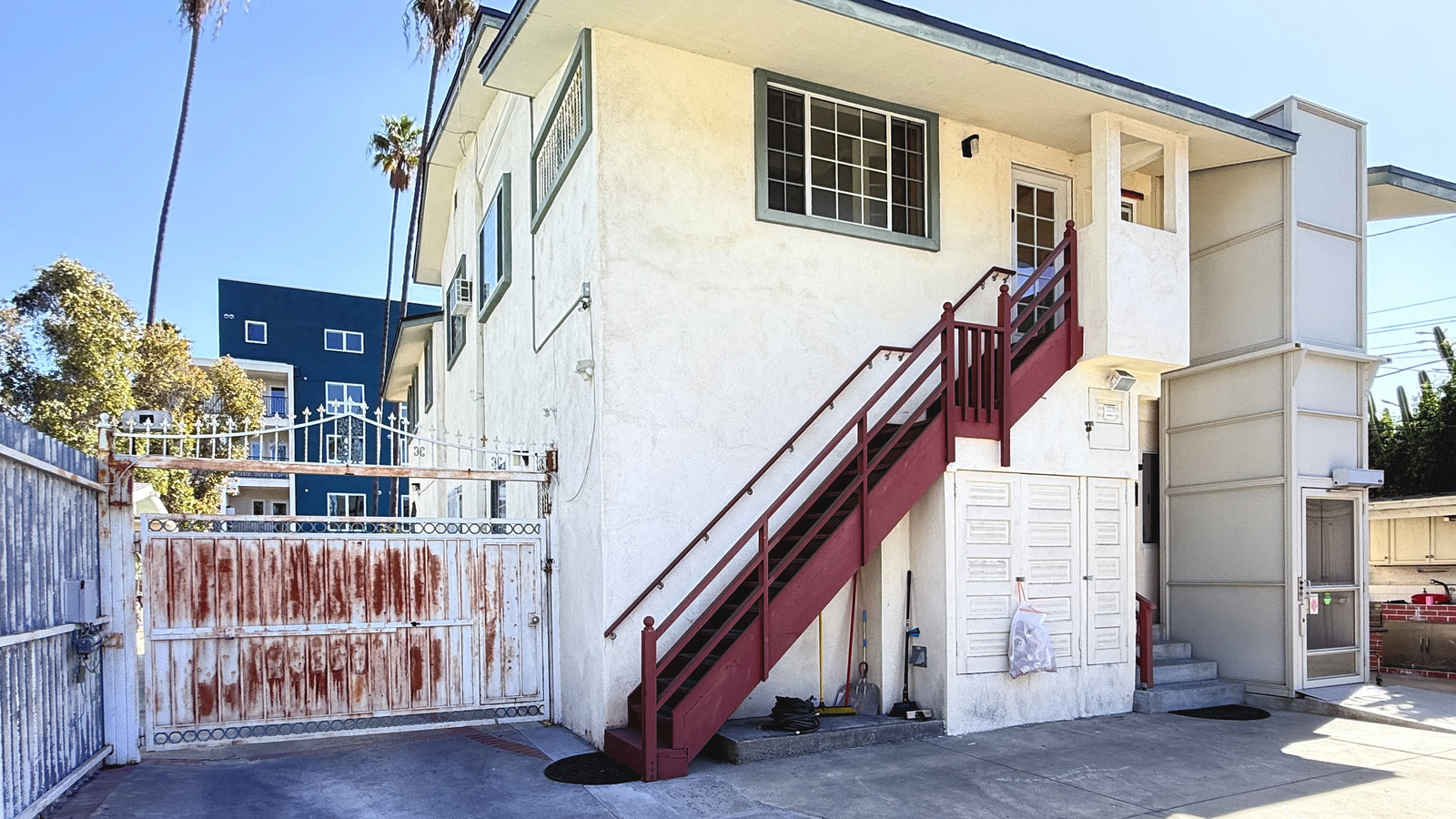 Rear elevation in sun: a red-painted exterior stair rising to an upper-level door, louvered cabinet doors beneath it, and the tall lift enclosure at right.