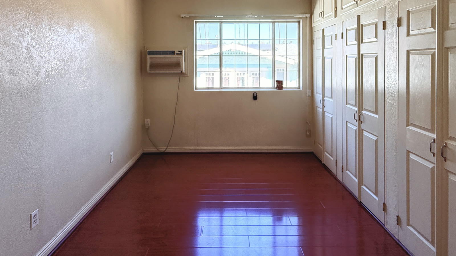Empty upstairs room with a grid window, a wall of white paneled closet doors with upper cabinets, and dark wood laminate flooring.