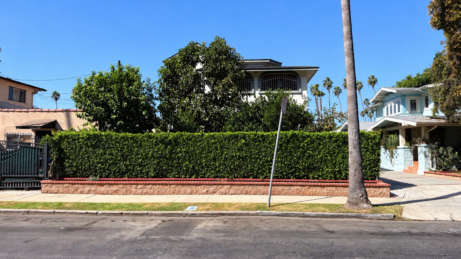 The house from across the street: a long clipped hedge on a brick-capped planter wall, mature trees behind it, and the arched, iron-grilled upper windows rising above, with palms along the block.